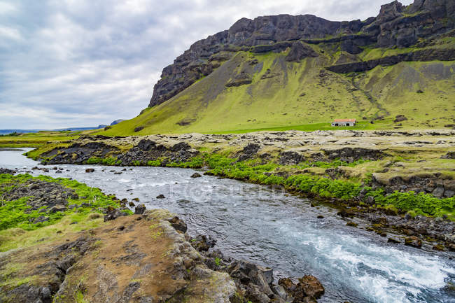Fiume blu dall'aspetto fresco corre lungo il bordo di una proprietà agricola con montagne vulcaniche sullo sfondo, Islanda — Foto stock