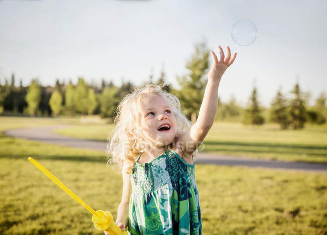 Linda chica joven soplando una burbuja y tratando de atraparlo en un parque - foto de stock