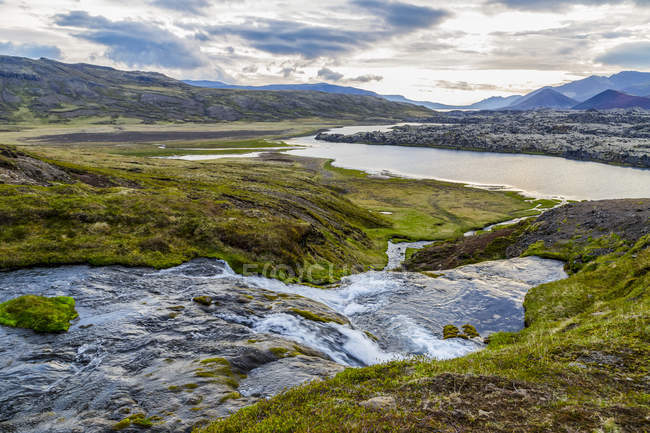 Pequeño río corre a través de un valle en el oeste de Islandia hacia el gran río de abajo y los lejanos picos volcánicos ricamente coloreados en la distancia, Islandia - foto de stock