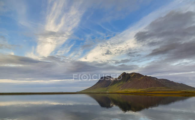 A rica terra vulcânica cria cores arrojadas na montanha contra o suave oceano azul e o reflexo do céu na Islândia Ocidental — Fotografia de Stock