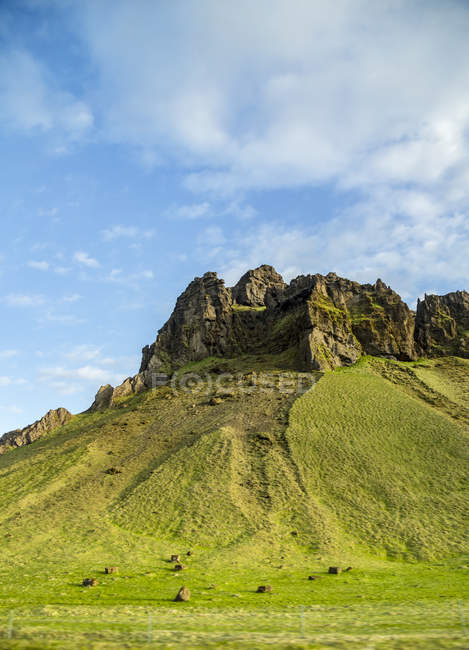 Escarpado pico rocoso que parece un monumento contra la ladera verde y el cielo azul, una vista común para ver en un viaje por carretera a través de Islandia, Islandia - foto de stock
