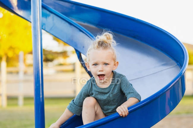 Young girl with blond hair playing in a playground and going down a slide — Stock Photo