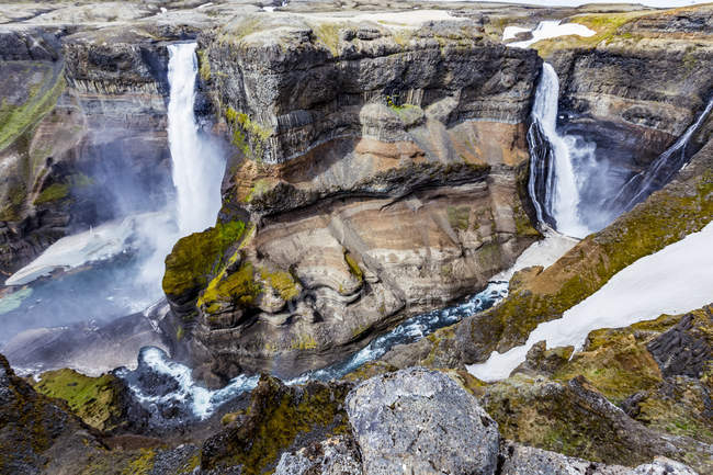 Los impresionantes colores de la tierra y las formaciones rocosas junto con la nieve sin derretir descansando en el fondo del valle de Haifoss entre dos cascadas, Islandia - foto de stock