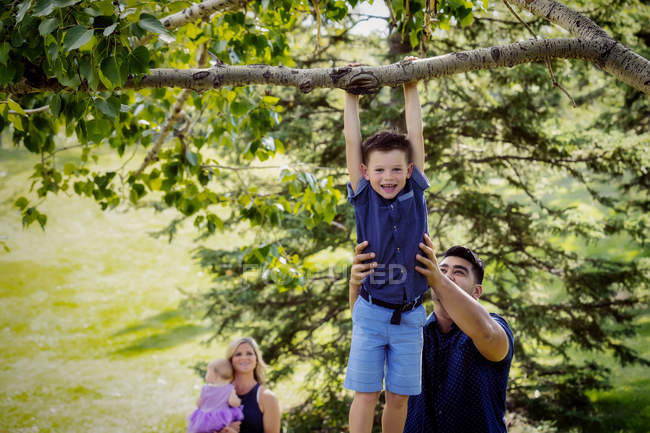 Una madre y un bebé vigilan mientras el padre sostiene a su hijo en lo alto para ayudarlo a colgar de una rama de árbol durante una excursión familiar en un parque en un cálido día de otoño - foto de stock