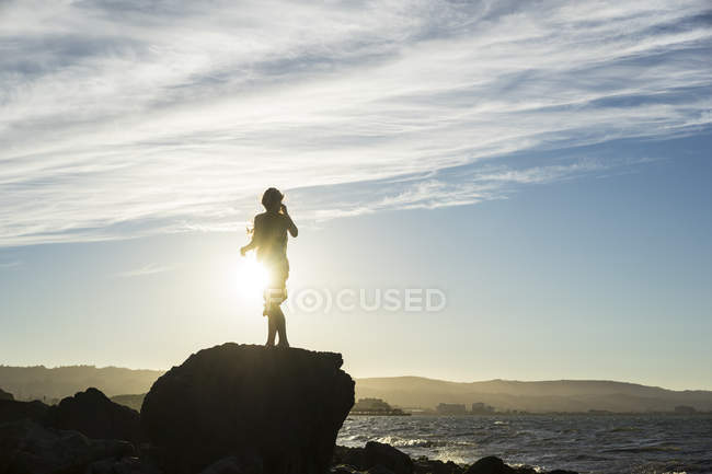 Una donna in piedi su una roccia che guarda fuori lungo la costa al tramonto, sagomata e retroilluminata dalla luce del sole; San Mateo, California, Stati Uniti d'America — Foto stock