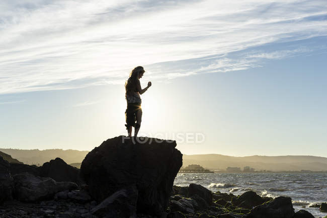 Une femme debout sur un rocher regardant le long de la côte au coucher du soleil, silhouette et rétroéclairé par la lumière du soleil ; San Mateo, Californie, États-Unis d'Amérique — Photo de stock