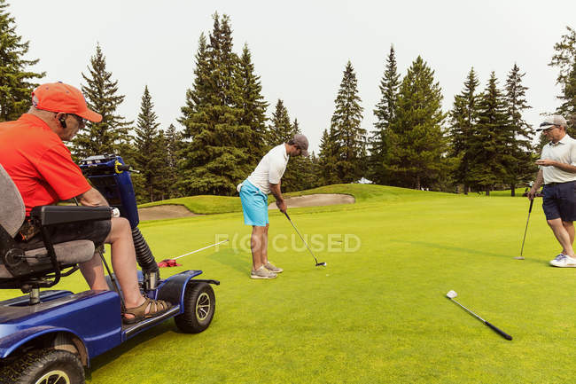 Deux golfeurs valides font équipe avec un golfeur handicapé utilisant un fauteuil roulant de golf motorisé spécialisé et se rassemblent sur un terrain de golf jouant la meilleure balle, Edmonton, Alberta, Canada — Photo de stock