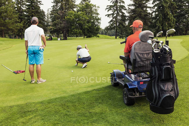 Due golfisti abili si uniscono con un golfista disabile usando una sedia a rotelle specializzata di golf motorizzata e mettendo insieme su un campo da golf verde che gioca la migliore palla, Edmonton, Alberta, Canada — Foto stock