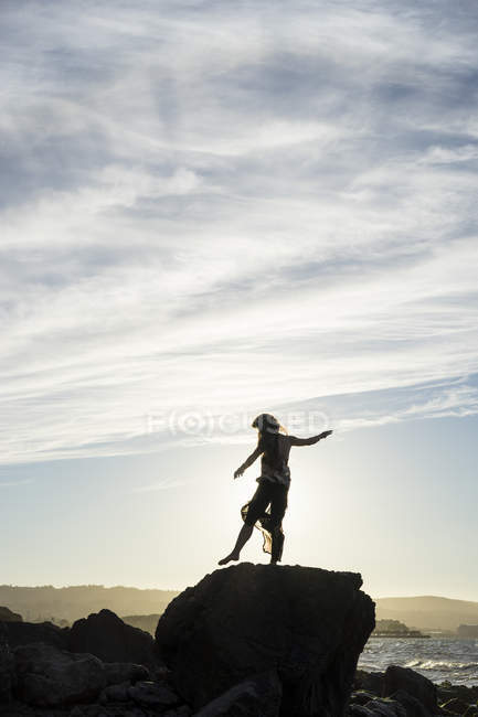 Una mujer de pie balanceándose sobre un pie en una roca con una vista de la costa al atardecer, silueta y retroiluminada por la luz del sol, San Mateo, California, Estados Unidos de América - foto de stock