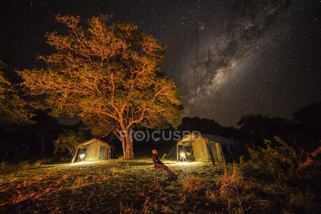 The milky way in the sky with a tents below in a bush camp as a man sitting looking up at the sky; Botswana — Foto stock
