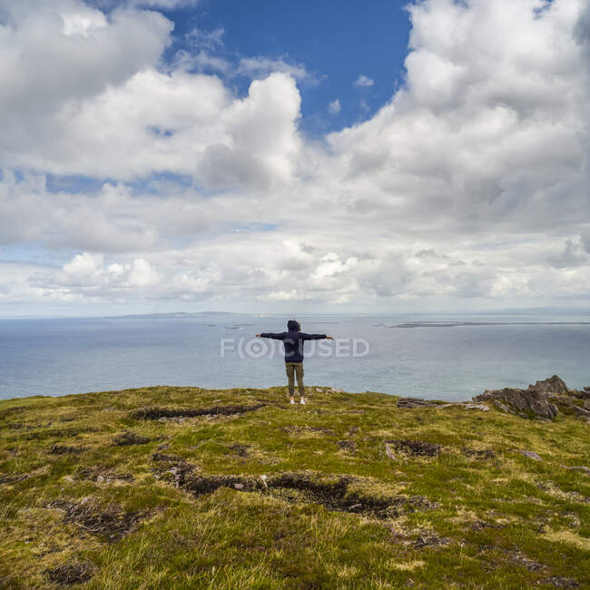 Mujer de pie con los brazos extendidos y mirando hacia el océano y la costa mientras caminaba en Brandon Point, Península de Dingle; Castlegregory, Condado de Kerry, Irlanda - foto de stock