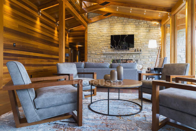 Sillas de madera marrón y gris tapizadas, mesa redonda y sofás con chimenea de piedra natural en la sala de estar de gran habitación - foto de stock