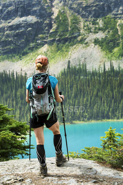 Female hiker standing on rocky cliff overlooking colorful alpine lake and mountain cliff in the background; British Columbia, Canada — Stock Photo