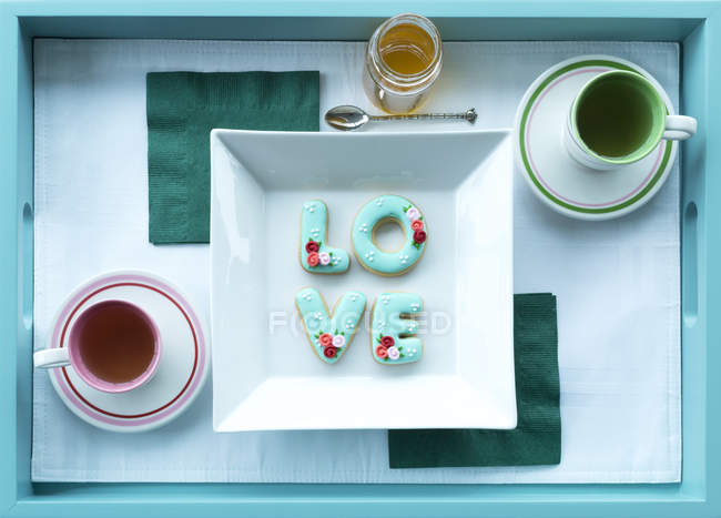 Tee in Tassen mit Untertassen und Keksen, die Liebe auf einem Tablett buchstabieren — Stockfoto