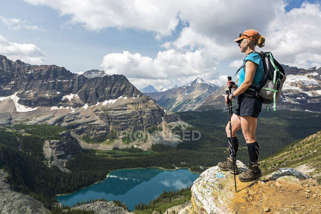 Senderista femenina de pie en el borde del acantilado con vistas a las montañas y el valle con lago alpino; Columbia Británica, Canadá - foto de stock