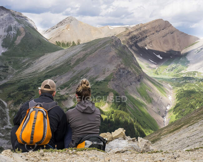 Male and female hikers sitting on a rock ridge overlooking a valley and mountain range in Kananaskis Country; Alberta, Canada — Stock Photo