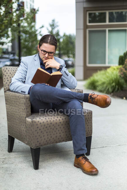 Молодий чоловік сидить, читаючи книжку в кріслі на відкритому подвір 