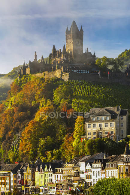Large medieval castle on top of a colourful treed hillside with fog, blue sky, clouds and village below; Cochem, Germany — Stock Photo