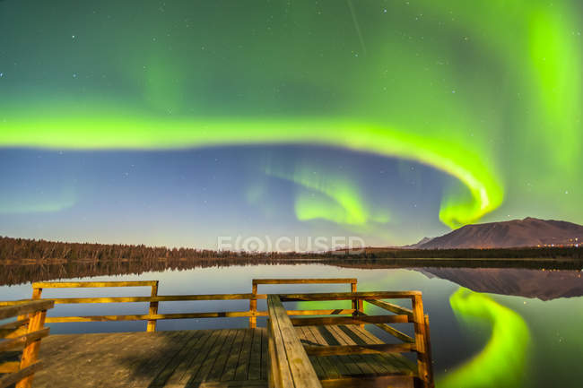 The Northern Lights are seen reflecting off Beach Lake on a clear, autumn night with a wooden dock in the foreground; Chugiak, Alaska, United States of America — Stock Photo