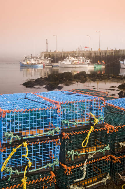 Casiers à homards empilés le long du rivage avec des bateaux attachés à un quai en arrière-plan, baie de Fundy ; Tiverton, Long Island, Nouvelle-Écosse, Canada — Photo de stock