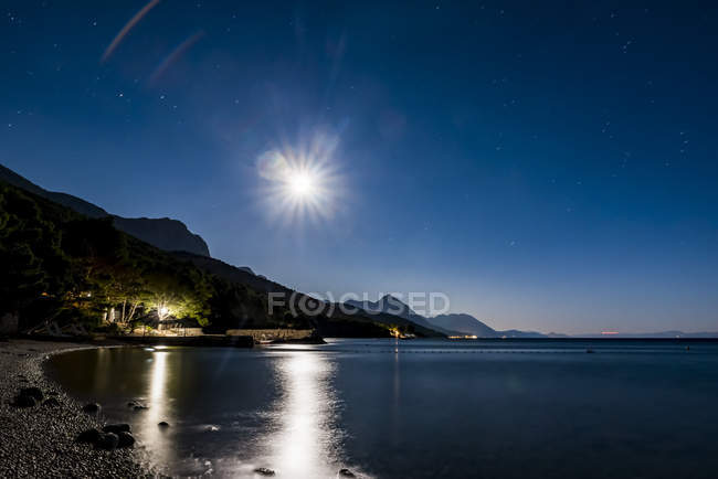 Makarska Riviera por la noche con la luz de la luna brillante que arroja luz sobre el agua tranquila a lo largo de la costa, Dalmacia, Croacia - foto de stock