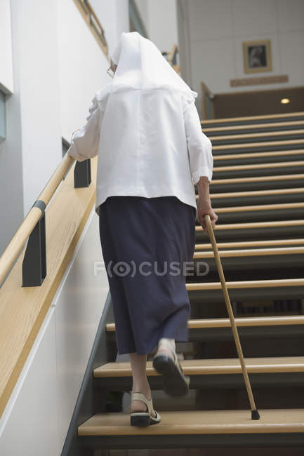 Вид сзади на монашку, поднимающуюся по лестнице с помощью трости — стоковое фото