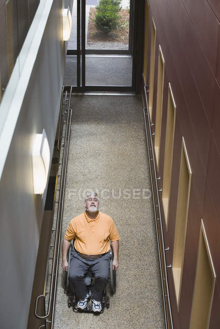 Высокий угол обзора мужчины средних лет с мышечной дистрофией, сидящего в инвалидном кресле в библиотеке — стоковое фото