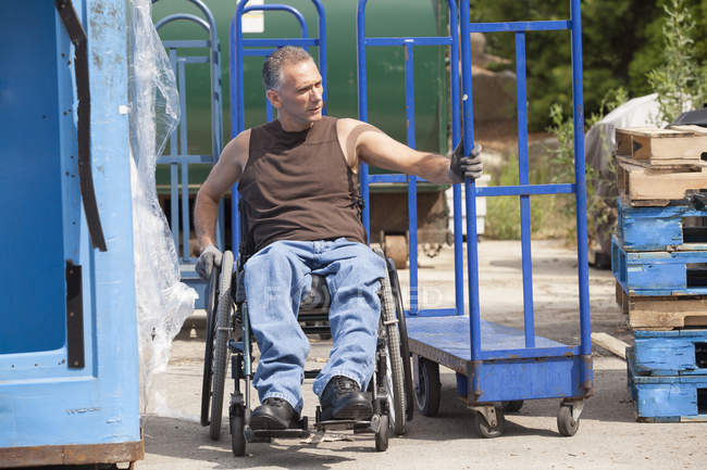 Cargando trabajador portuario con lesión medular en una silla de ruedas moviendo un camión de mano - foto de stock
