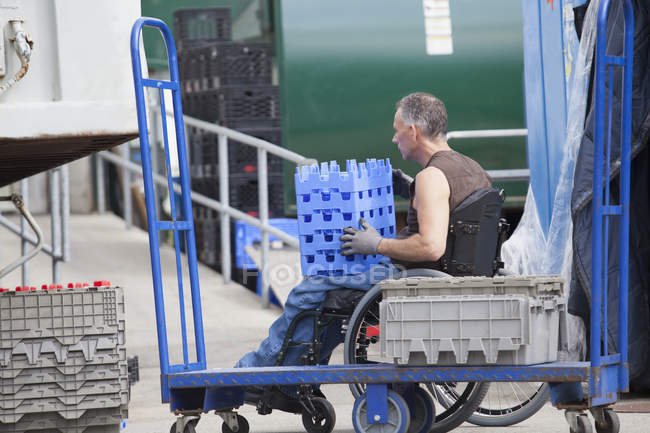 Trabajador portuario de carga con lesión medular en una silla de ruedas moviendo bandejas de inventario apiladas - foto de stock