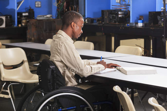 Étudiant en fauteuil roulant prenant des notes tout en lisant le manuel dans la salle de classe CVC — Photo de stock