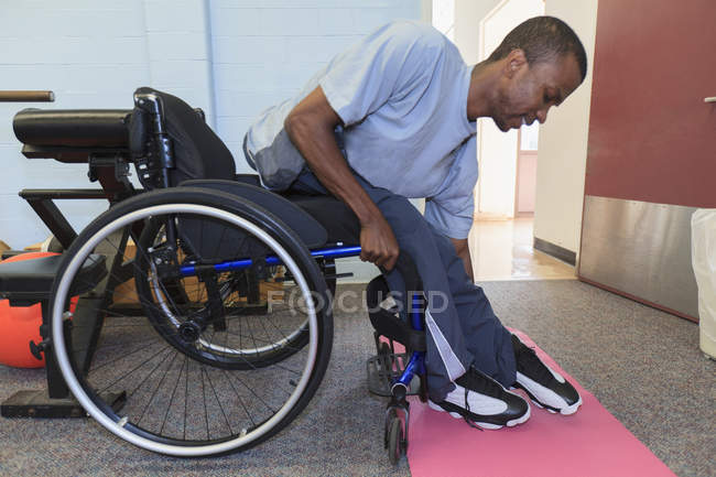 Hombre que tenía meningitis espinal subiéndose a su silla de ruedas desde una almohadilla de yoga - foto de stock
