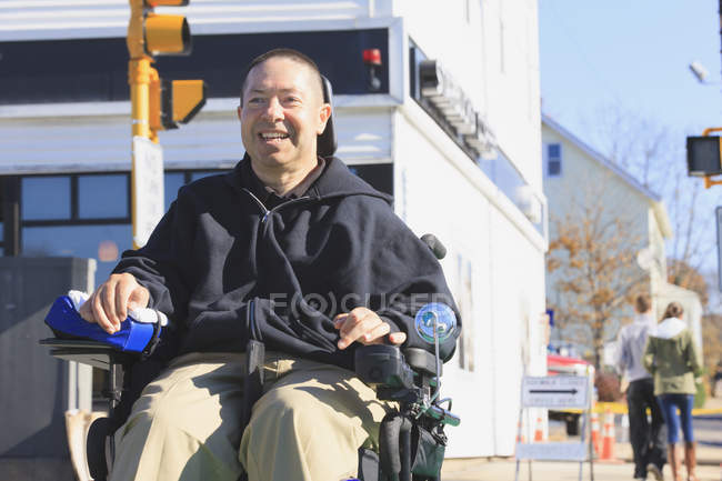 Hombre con lesión medular y brazo con daño nervioso en silla de ruedas motorizada cruzando calle pública mientras hace compras - foto de stock