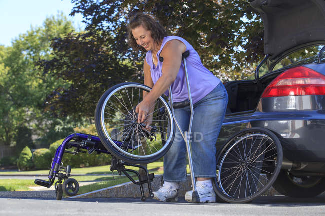 Женщина со Спиной Бифидой с помощью костылей разбирает инвалидное кресло на части для передвижения в машине — стоковое фото