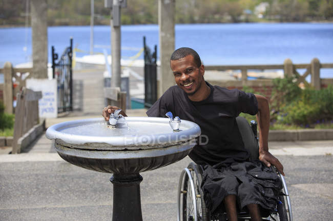 Homem que teve meningite espinhal numa cadeira de rodas a beber de uma fonte — Fotografia de Stock
