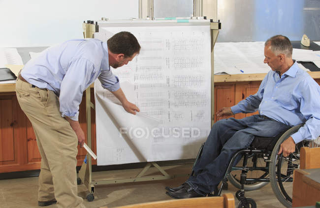 Dos ingenieros de proyecto que utilizan un bastidor colgante para los planes del sitio de trabajo, uno en una silla de ruedas con una lesión en la médula espinal - foto de stock