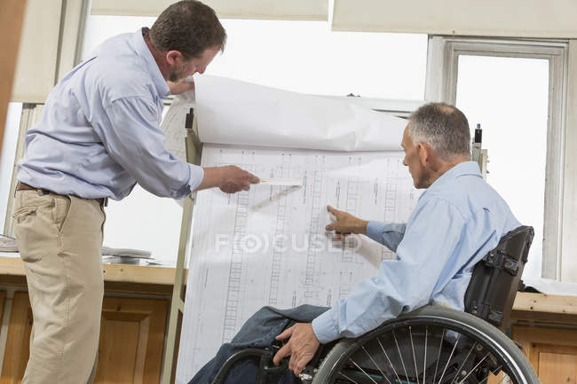 Deux ingénieurs de projet utilisant un support suspendu pour les plans de chantier, un dans un fauteuil roulant avec une blessure à la moelle épinière — Photo de stock