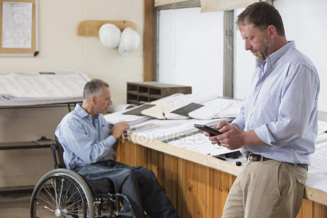 Zwei Projektingenieure überprüfen mit ihren Tablets die Baustellenpläne, einer sitzt im Rollstuhl und ist querschnittsgelähmt — Stockfoto