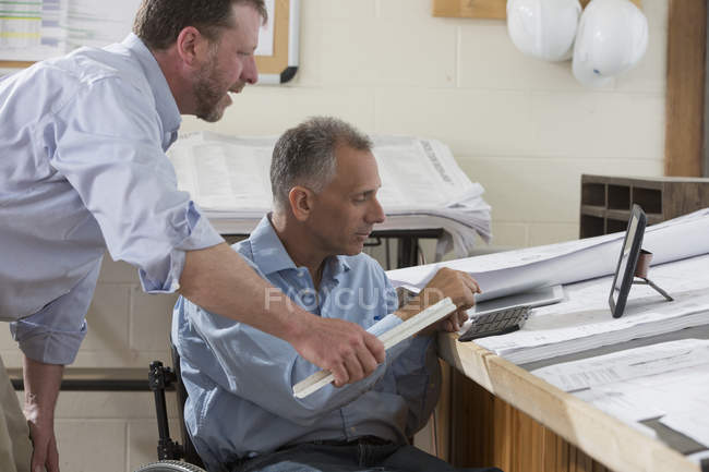 Zwei Projektingenieure überprüfen mit ihren Tablets die Baustellenpläne, einer sitzt im Rollstuhl und ist querschnittsgelähmt — Stockfoto