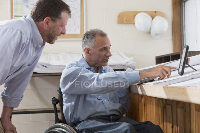 Два проектних інженери використовують планшет, щоб перевірити плани на місці роботи, один у інвалідному візку з Spinal Cord Injury — стокове фото