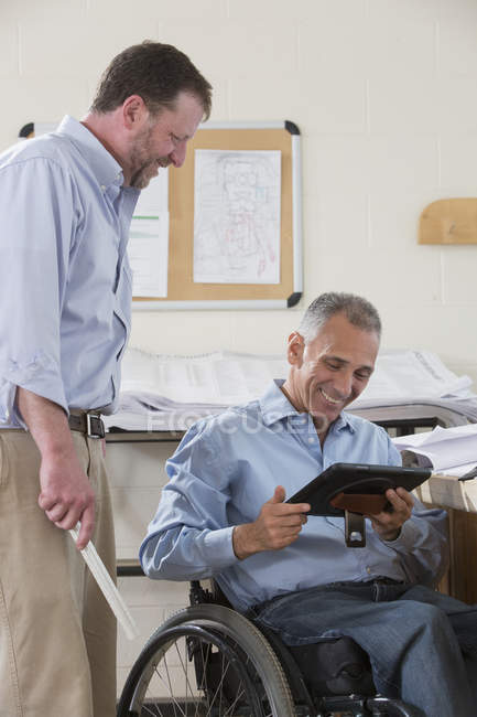 Dois engenheiros de projeto usando seu tablet para verificar os planos do local de trabalho, um em uma cadeira de rodas com uma lesão na coluna vertebral — Fotografia de Stock