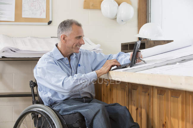 Инженер-проектировщик с помощью планшета проверяет планы места работы, находясь в инвалидном кресле с травмой спинного мозга — стоковое фото