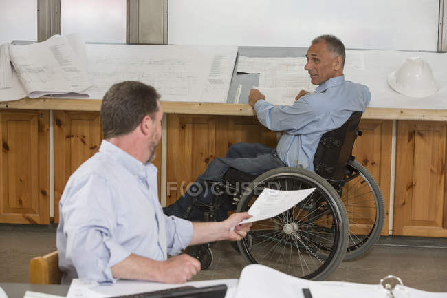 Dois engenheiros de projeto fazendo papelada sobre o trabalho, um em uma cadeira de rodas com uma lesão na medula espinhal — Fotografia de Stock