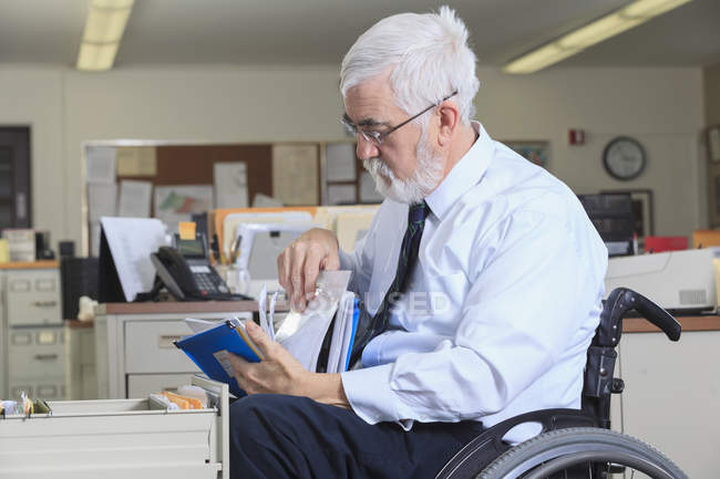 Uomo con distrofia muscolare su una sedia a rotelle cercando scartoffie nel cassetto del suo ufficio — Foto stock