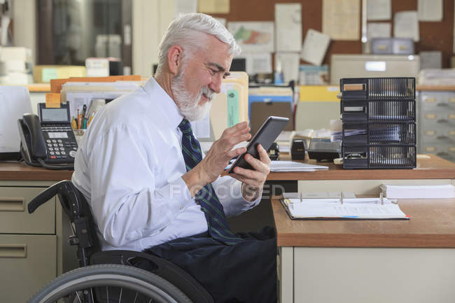 Uomo con distrofia muscolare in sedia a rotelle utilizzando un tablet alla scrivania del suo ufficio — Foto stock
