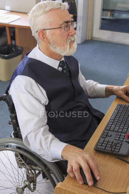 Человек с мышечной дистрофией в инвалидной коляске, работающий за компьютером в офисе — стоковое фото