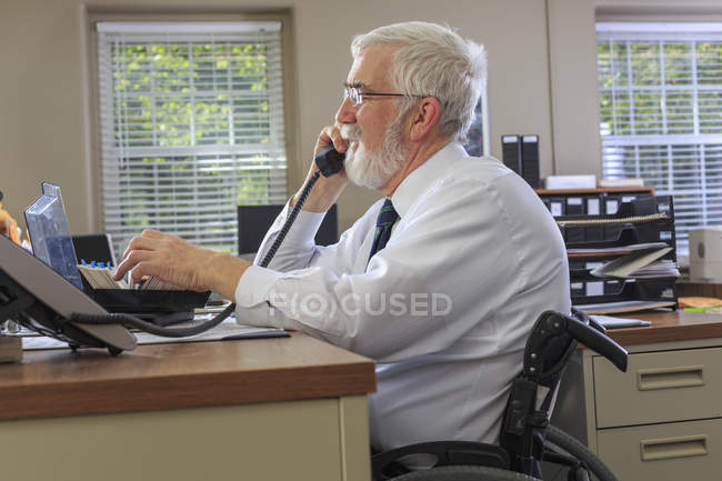 Человек с мышечной дистрофией в инвалидном кресле разговаривает по телефону в офисе — стоковое фото