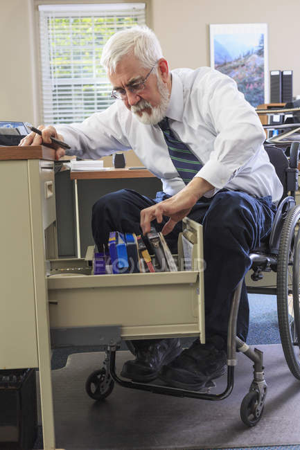 Man with Muscular Dystrophy in a wheelchair taking files of information from his desk drawer — Stock Photo