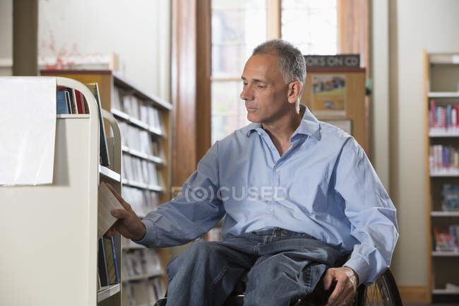Uomo su una sedia a rotelle con una lesione al midollo spinale che organizza libri in una biblioteca — Foto stock