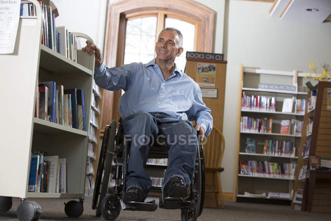 Un homme en fauteuil roulant blessé à la moelle épinière déplaçant une étagère de livres dans une bibliothèque — Photo de stock