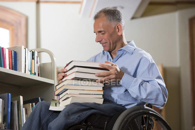 Man in a wheelchair with a Spinal Cord Injury choosing books from a shelve in a library — Stock Photo